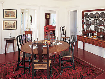 The red paint of the floor against the white walls of the dining room seems to elevate the dining table and chairs. The English dresser was acquired in Petworth, England, and holds some of the couple's collection of English pewter. It also holds a sixteenth-century ceramic horse and a coin bank thought to be Chinese. Two nineteenth-century English prints, one of which depicts the Royal Mail, and the other, the Birmingham Tally Ho, hang above one of a pair of Queen Anne chairs and a fine splay leg stand. The living room, with the Silas Hoadley tall clock, is seen in the room beyond. In the front entry, a William & Mary dressing table is placed below a Chippendale looking glass.