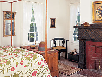 A prized coverlet on a tester bed in an upstairs bedroom complements the colors on the facade of a carved and painted Connecticut chest nearby, purchased from dealer David Schorsch. The cats of the hooked rug play off the carved lamb placed on a small early eighteenth-century green storage box on ball feet and with snipe hinges. The decorative marbleizing of the fireplace surround was completed after the house was built. The walls are hung with a  portrait of a young lady by itinerant artist William Matthew Prior (1806-1873), and an eglomisé gilt mirror, both from the nineteenth century.