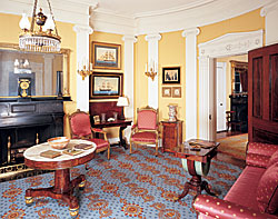 Click Here for Enlarged View Classical themes prevail in the reception room with scrolling volutes atop Ionic pilasters and a lotus and palmette pattern carved into the architrave above the doorway. Evidence of later gasification and electrification has been removed from the chandelier (Johnston-Brooks of Birmingham, England, circa 1820), so it now appears as it would have when burning oil. The carved mahogany work table with demi-lune bag, raised on acanthus carved saber legs with brass toe caps and casters was made in Boston, probably by Timothy Hunt. The marble-top center table, 1825-1830, maker unknown, was made in New York.